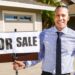 Smiling real estate agent with a for sale sign in front of a house