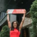 Smiling woman holding a 'Sold' sign outside a building, celebrating a successful sale.